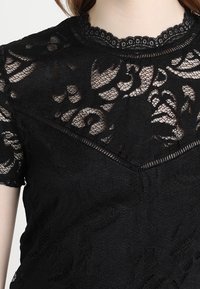 Černý krajkový top s krátkými rukávy, zdobený květinovými vzory, texturovanými látkami, vlnitým výstřihem a dekorativním lemováním.