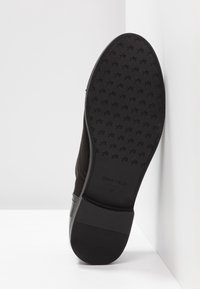 Semelle d'une chaussure de ville noire avec une semelle à motif, la marque "ANNA FIELD" et la taille 37 visibles, reposant contre une surface blanche.
