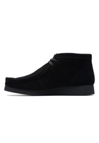 Bota de ante negra hasta el tobillo con punta redonda, detalles de costura y suela de goma baja. Presenta un único lazo decorativo en la parte frontal.