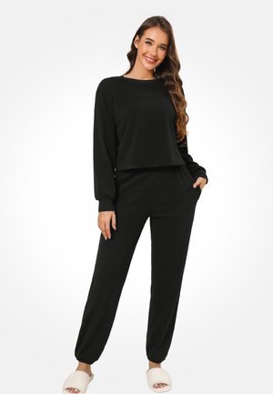 Glunderende vrouw met lang golvend haar, gekleed in een zwarte top met lange mouwen en bijpassende broek, en witte, pluizige pantoffels, poserend met één hand in de zak.