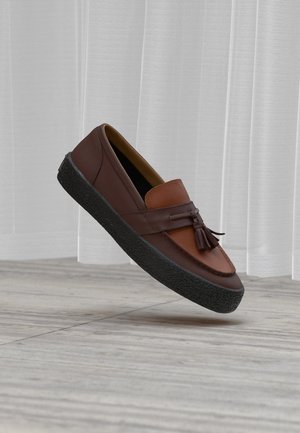 Barna bőrből készült mokaszin bojttal és vastag fekete gumitalppal, amely egy világos fadézsa fölött lebeg, fehér függönyök előtt.