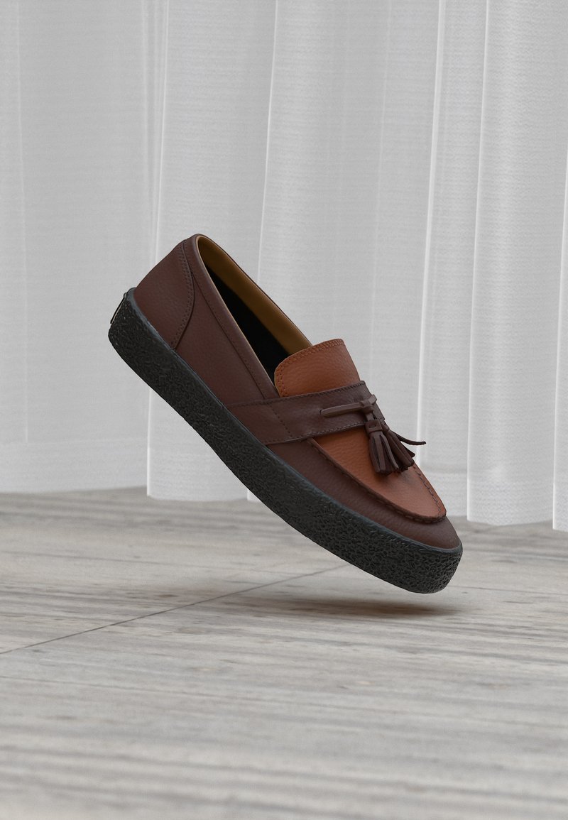 Mocassin en cuir marron avec pompons et semelle épaisse en caoutchouc noir flottant au-dessus d'un sol en bois clair devant des rideaux blancs.