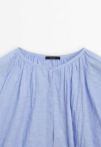 Lys blå bluse med rund halsudskæring, plisseret design og blødt, tekstureret stof. Indeholder et sort mærke ved halsen med brandnavn.