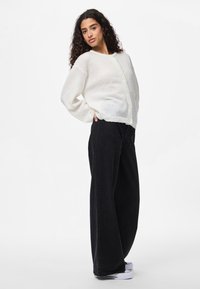 Cardigan blanc en maille avec manches longues et encolure ronde, présentant une texture douce et une fermeture à boutons. Associé à un pantalon noir large.