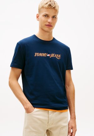 Giovane uomo con capelli biondi corti che indossa una maglietta blu navy "Tommy Jeans" e pantaloni beige, in piedi con una mano in tasca.