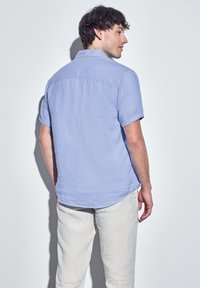 Camicia azzurra a maniche corte con collo, tessuto testurizzato e orlo curvo. Abbinata a pantaloni di lino di colore chiaro.