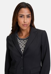 Černý úzký blazer se dvěma knoflíky a bočními kapsami, nošený přes vzorovaný černobílý top s kulatým výstřihem.