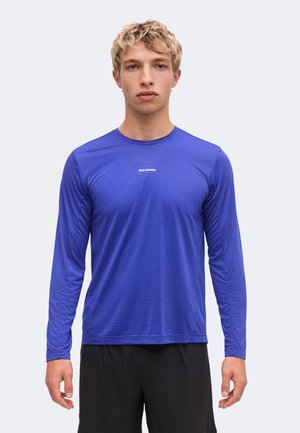 Joven con cabello rubio corto, vistiendo una camiseta azul de manga larga Salomon y pantalones cortos negros, de pie frente a un fondo liso.