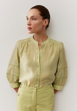 Frau mit zurückgekämmtem Haar, die eine hellgrüne, geknöpfte Bluse und passende hochtaillierte Hose trägt und gegen eine schlichte Wand nach links schaut.