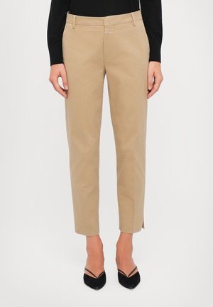 Femme portant un pantalon beige raccourci avec des fentes latérales, associé à des escarpins noirs à bouts pointus et un haut noir à manches longues.