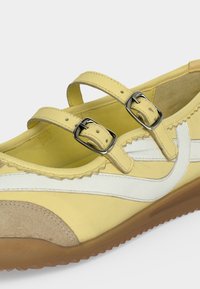 Chaussure Mary Jane jaune avec deux brides réglables à boucle, détail latéral blanc, bout beige et semelle texturée marron.