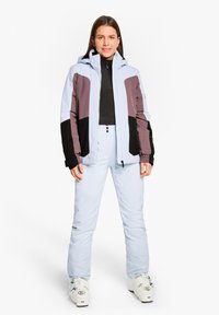 Abbigliamento da sci con una giacca azzurro chiaro con accenti viola scuro e neri, abbinata a pantaloni azzurro chiaro e scarponi da sci bianchi.