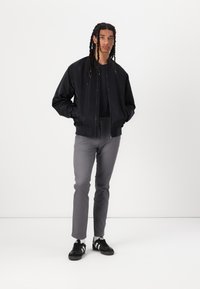 Chaqueta bomber negra con cierre frontal, combinada con pantalones gris entallados y zapatillas negras. Tela suave, diseño informal, de pie sobre un fondo blanco.