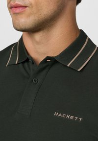 Man met een donkergroene polo met twee beige strepen op de kraag en "HACKETT" geborduurd op de borst.