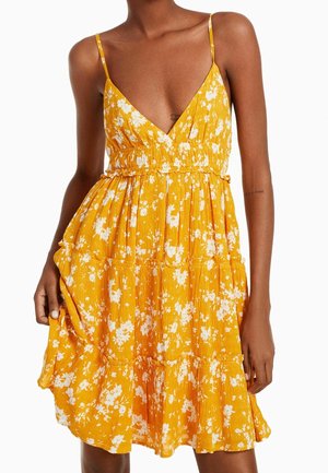 Bezrekawna żółta sukienka letnia z białym kwiatowym wzorem, dekoltem w kształcie litery V, marszczoną talią i warstwową spódnicą, noszona przez kobietę.