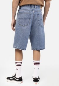 Osoba koja nosi svijetloplave traper kratke hlače, bijele čarape s bordo prugama, crne tenisice i smeđu majicu, stojeći okrenuta leđima prema kameri.