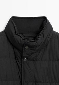 Black padded vest with a high collar, featuring snap buttons and a smooth texture. The fabric has a subtle sheen and horizontal stitching.