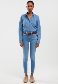 Mlada žena stoji s neutralnim izrazom lica, odjevena u svijetloplavu traper košulju i traperice, smeđi remen i bež tenisice, na jednobojnoj pozadini.