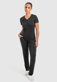 Zwarte V-hals t-shirt gecombineerd met zwarte broek, met een soepele textuur en een ontspannen pasvorm. Model staat met handen in de zakken, draagt witte schoenen.