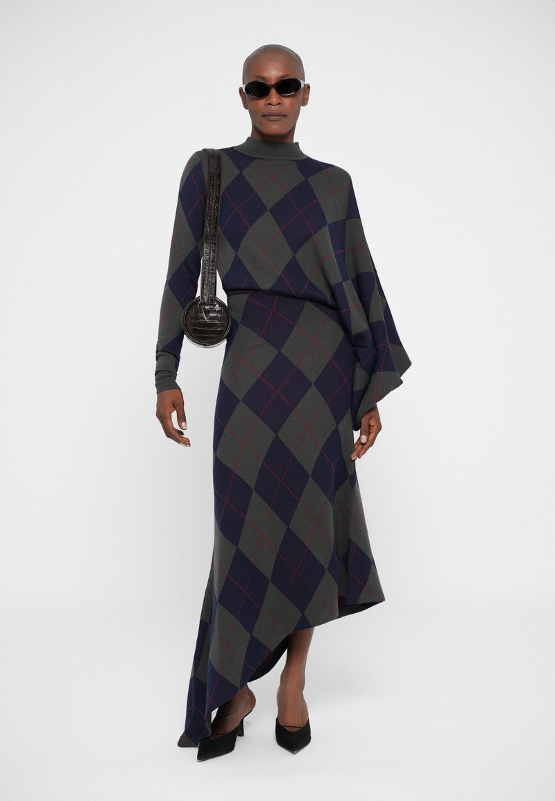 Langmouwige jurk met een hoge hals, voorzien van een groen, marineblauw en rood geruit patroon. Geaccentueerd met een zwarte handtas en puntige hakschoenen.
