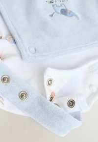 Close-up van lichtblauwe en witte babykleding met metalen drukknoopjes en een kleine geborduurde blauwe walvisdetail.