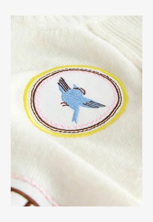 Weißer Stoff mit einem kreisförmigen, bestickten Patch, der einen blauen Vogel zeigt, umrandet von Gelb mit rosa Stickerei, was Textur und Farbkontrast hervorhebt.