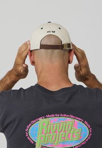 Béžová čepice s tmavě zeleným knoflíkem a nápisem „est. '88“ na zadní straně, upevněná hnědým koženým páskem. Tmavé tričko s barevnou grafikou.