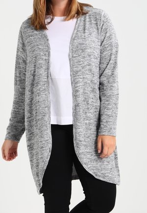 Žena oblékající dlouhý šedý kardigan s otevřeným předním dílem přes bílou košili a černé kalhoty, stojící před neutrálním pozadím.