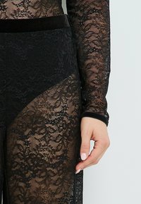Bodysuit en dentelle noire avec manches longues, motifs floraux transparents, une taille en velours et une finition lisse.
