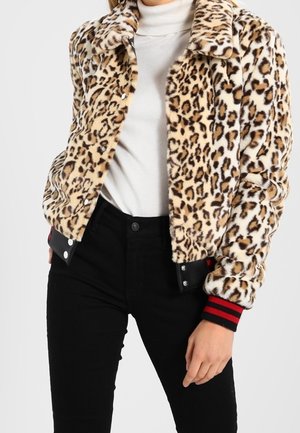Vrouw draagt een faux fur jas met luipaardprint met zwarte en rode gestreepte manchetten over een witte coltrui en zwarte broek.