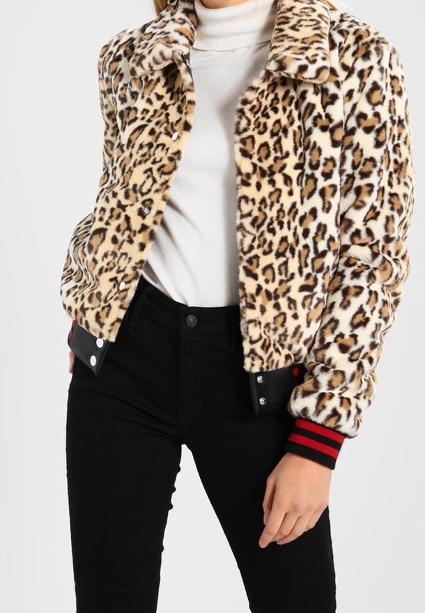 Femme portant une veste en fausse fourrure imprimé léopard avec des poignets rayés noirs et rouges, par-dessus un col roulé blanc et un pantalon noir.