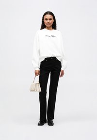 Tricou alb cu logo-ul "Tommy Hilfiger", pantaloni negri mulati și o poșetă crem. Pantofi negri completează ținuta. Fundal simplu.