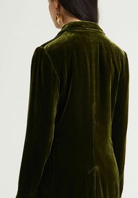 Veste en velours vert à coupe ajustée, col cranté et manches longues. Texture lisse avec un léger éclat. Dispose d'une fente à l'arrière.