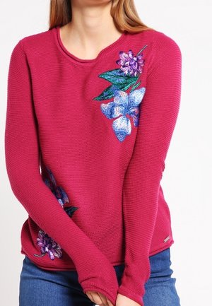 Femme portant un pull moulant texturé rose foncé avec une broderie florale violette et bleue sur la poitrine et le côté inférieur, assorti à un jean bleu.