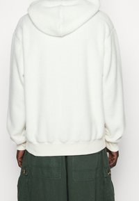 Weißer Fleece-Kapuzenpullover mit weicher Textur, abgerundeter Kapuze sowie gerippten Bündchen an Ärmeln und Saum. Getragen über grüne Cargohosen mit Seitentaschen.