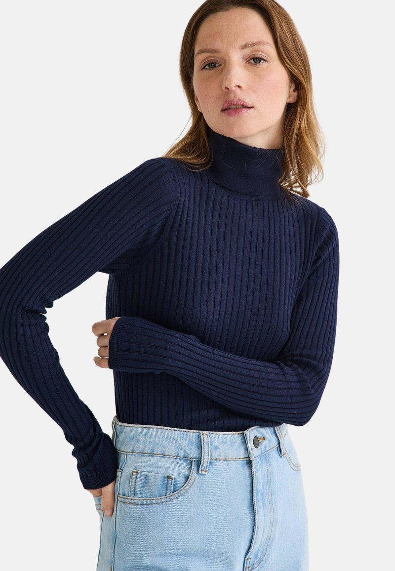 Col roulé côtelé bleu marine en maille, avec des manches longues et une coupe ajustée. Associé à un jean taille haute bleu clair.