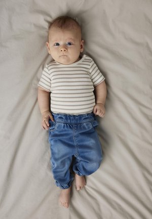 Juostuota smėlio ir baltos spalvos medvilninė trumpomis rankovėmis marškiniai derinami su mėlynais džinsiniais elastingais kelnais ant šviesiai smėlio fono.