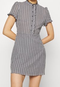 Schwarz-weißer Karo-Kleid mit kurzen Rüschenärmeln, hohem Dekolleté und vorderem Knopfverschluss; tailliert und mit weit auslaufendem Rock.