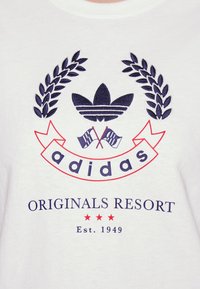 Witte t-shirt met blauw geborduurd logo met drie bladeren, "adidas," en "ORIGINALS RESORT" tekst in rood en blauw, met drie rode sterren.