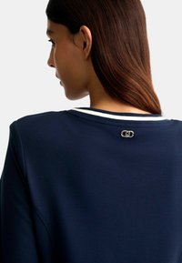Maglietta girocollo blu navy con colletto a righe bianche, dotata di una chiusura metallica decorativa sul retro. Tessuto dalla consistenza liscia.