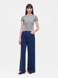 Femme debout portant un t-shirt gris ajusté, un jean large bleu foncé et des chaussures marron, une main dans la poche, sur fond uni.