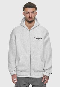 Haut à zip gris avec un logo sur le devant, en tissu doux, poches avant et poignets côtelés. Texture légère et coupe décontractée.