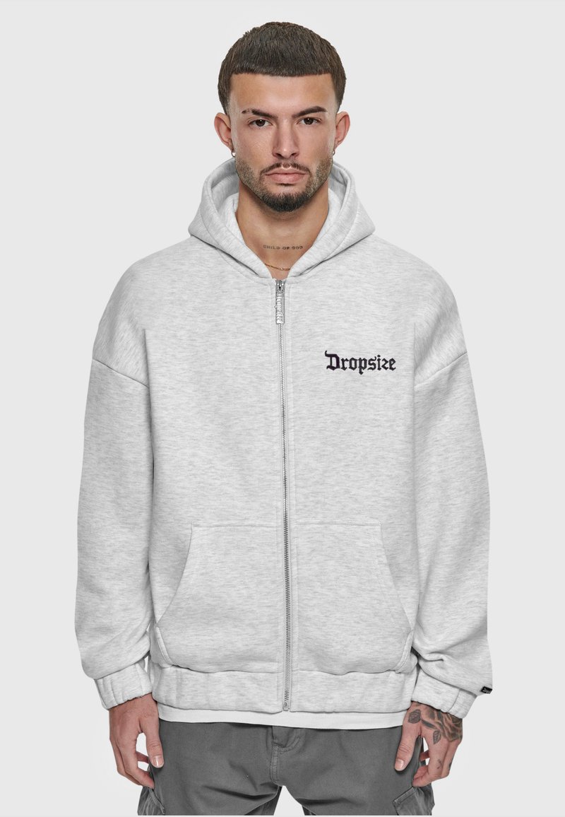 Haut à zip gris avec un logo sur le devant, en tissu doux, poches avant et poignets côtelés. Texture légère et coupe décontractée.