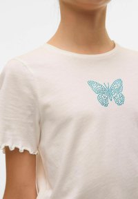 T-shirt en coton crème avec un design de papillon orné de pierres turquoise, manches courtes avec des bords festonnés, texture douce.
