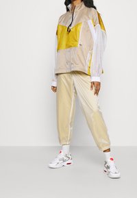 Beige och gul meshtröja med hög krage, lätt tyg och elastiska ärmslut, i kombination med glänsande beige joggers och vita sneakers.