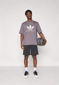 Pelēks pārliels T-krekls ar baltu uzrakstu "BASKETBALL", kopā ar melniem šortiem un baltām sporta kurpēm. Rokās tur basketbola bumbu.