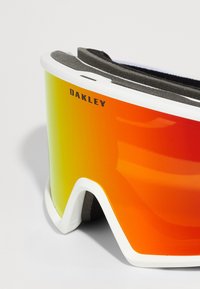 Lunettes de ski Oakley avec une monture blanche, un verre miroir dégradé orange au jaune et un rembourrage en mousse noire pour plus de confort.