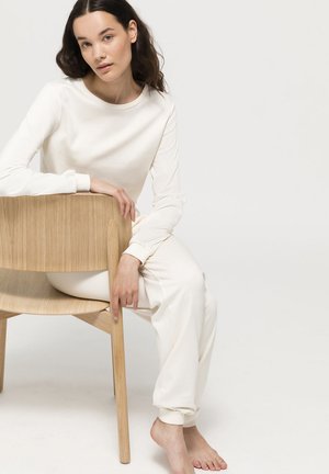 Helles beiges Loungewear-Set aus weichem Stoff, bestehend aus einem Langarmoberteil und tapered Hosen, kombiniert mit einem Holzstuhl.