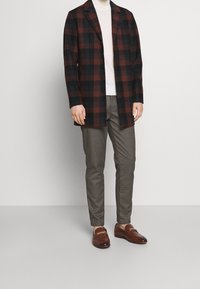 Hnědo-černá kostkovaná bunda, zapnutá, v kombinaci s světlým svetrem a kostkovanými kalhotami. Hnědé mokasíny se zlatými kovovými akcenty.