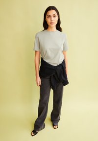 Helles graues T-Shirt mit kurzen Ärmeln, schwarzer Pullover um die Taille gebunden, dunkelgraue Schlagjeans, offene Sandalen mit Absatz, stehend vor einem hellgrünen Hintergrund.